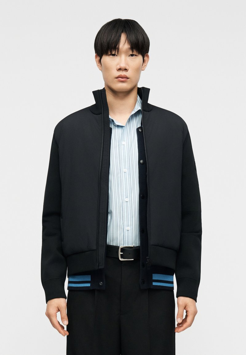 Νέος άνδρας με μαύρο μπουφάν με φερμουάρ πάνω από ανοιχτό μπλε πουκάμισο με ρίγες και μαύρο παντελόνι με μαύρη ζώνη, στέκεται μπροστά σε απλό φόντο.