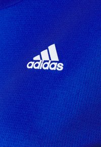 Sininen urheilukangas, jossa on teksturoitu kuvio, vasemmassa yläkulmassa valkoinen Adidas-logo ja kolme valkoista raitaa.