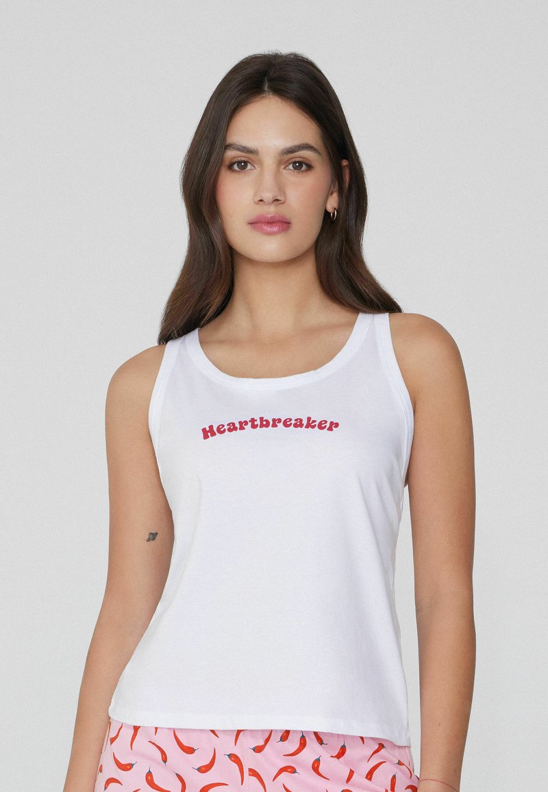 Top de tirantes de algodón blanco que presenta el texto "Heartbreaker" en rojo, con un corte holgado y tirantes anchos, combinado con un pantalón de fondo estampado en rosa.