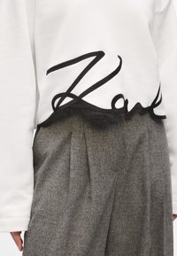 White sweatshirt featuring a black embroidered cursive word along the bottom hem, paired with gray pleated wool trousers.