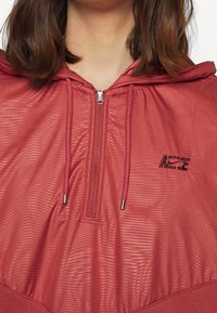 Červená lehká bunda s kapucí, zipovým uzávěrem, texturovanou látkou s horizontálními pruhy a malým logem Nike na hrudi.