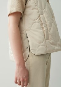Beige gesteppte Weste mit abgerundeten Formen, seitlichen Druckknopfdetails und einer Brusttasche, getragen über einem hellen Pullover und einer Hose.