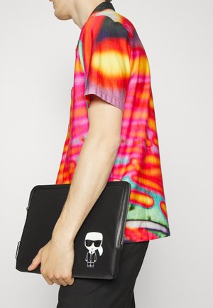 Housse de portable zippée noire présentant un graphique de dessin animé d'une figure portant des lunettes de soleil, tenue par une personne vêtue d'une chemise colorée et psychédélique.