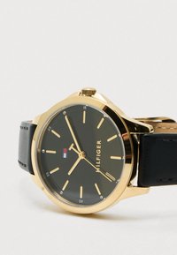 Guldtonet ur med en sort urskive, sølv time markører og et sort læderarmbånd. Har et logo i rød, hvid og blå. Klassisk rund form.