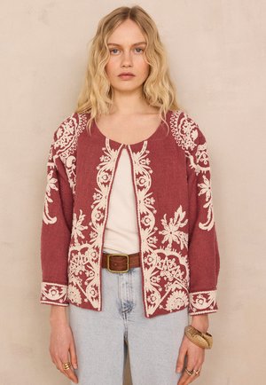 Femme aux longs cheveux blonds porte un cardigan bordeaux avec une broderie florale blanche, un haut blanc, un jean bleu clair et une ceinture marron.