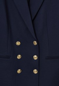 Blazer blu navy con un design strutturato, fronte doppiopetto e sei bottoni dorati. Presenta doppie tasche e una texture di tessuto liscia.