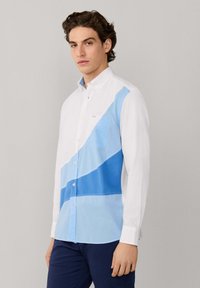 Jonge man met een wit overhemd met diagonale licht- en donkerblauwe kleurvlakken, staand tegen een effen grijze achtergrond.