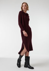 Bordeaux ribbelgebreide jurk met een colhals en zijsplitten, gecombineerd met zwarte enkellaarsjes met klapaccenten. Glad textuur.