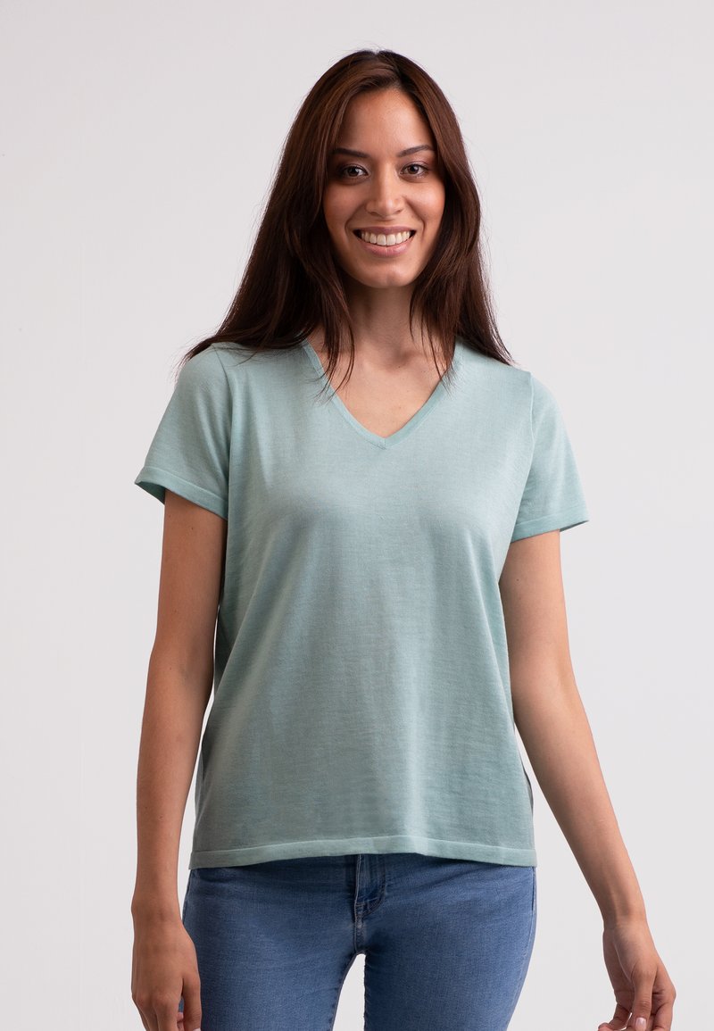 Svetle zelené tričko s krátkým rukávem a výstřihem do V vyrobené z měkké a hladké tkaniny, s volným střihem, spárované s modrými džíny na neutrálním pozadí.