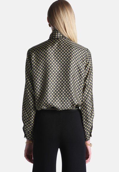 Femme aux cheveux blonds raides porte un chemisier noir à manches longues avec un motif géométrique doré et un pantalon noir, vue de dos.