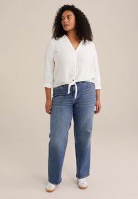 Camisa de algodón blanca con botones y nudo en el frente, combinada con jeans de mezclilla azul de corte relajado y zapatillas blancas. Textura suave y ajuste casual.