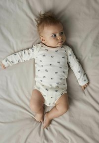 Langarm-Babystrampler in Creme mit einem verspielten Muster aus blauen Traktoren, weichem Stoff und Druckknopfverschluss an der Unterseite für einen einfachen Wechsel.