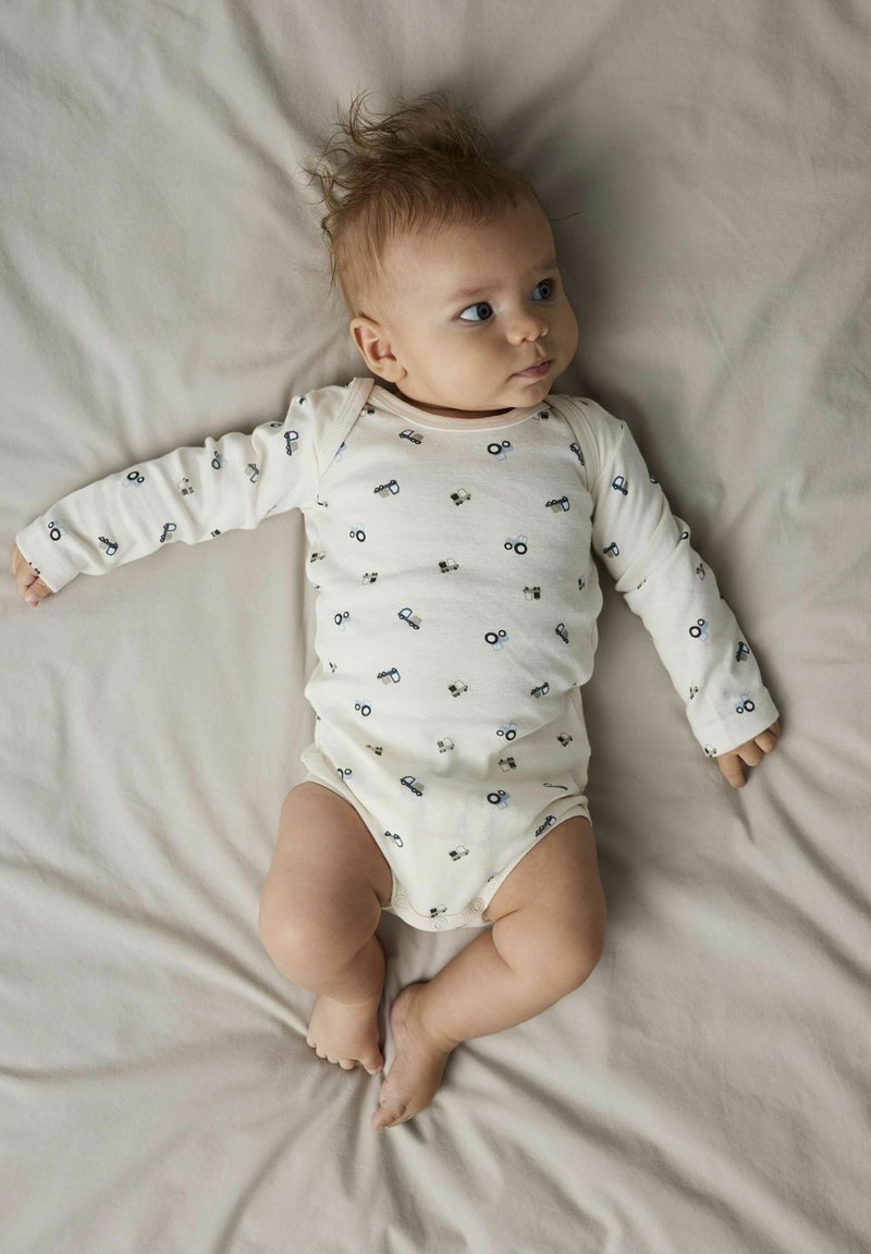 Langarm-Babystrampler in Creme mit einem verspielten Muster aus blauen Traktoren, weichem Stoff und Druckknopfverschluss an der Unterseite für einen einfachen Wechsel.