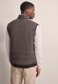 Grau gestepptes Puffer-Weste mit Fischgrätmuster, mit Kragen und dunklem braunen Besatz. Über einem weißen gerippten Pullover und hellbeigen Hosen getragen.