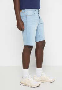 Lichtblauwe denimshorts met een rechte pasvorm, vijf zakken en subtiele stiksels, gedragen met witte sportschoenen en sokken.