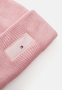 Bonnet tricoté rose avec un motif texturé et une étiquette logo rectangulaire affichant le nom de la marque et un emblème tricolore en rouge, blanc et bleu.