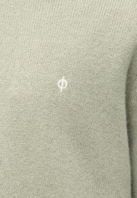 Tecido de malha verde claro com pequeno logotipo circular bordado em branco centrado na área do peito.