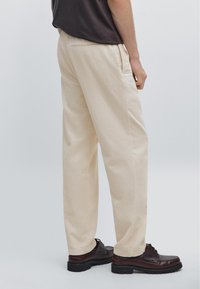 Person wearing beige corduroy pants, dark brown shoes, and a dark top, shown from mid-back to feet against a plain background.