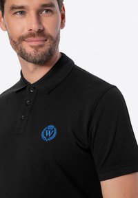 Černé polo tričko s límečkem, knoflíkovou légou a modrou vyšívanou logem se vzorem koruny a vavřínových listů. Hladká textilie.