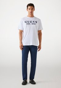 Weißes Baumwoll-T-Shirt mit navyblauer "GUESS EST. 1981" Druck; kombiniert mit dunkelblauen Jeans und schwarzen Slippern. Figurbetonter, lässiger Look.