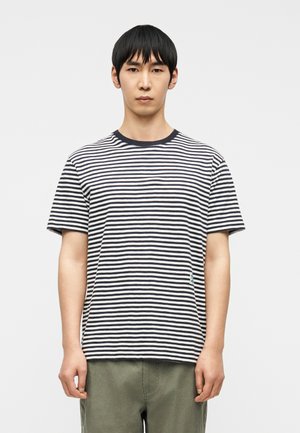 Muž v čierno-bielom tričku s horizontálnymi pruhmi a olivovo zelených nohaviciach, stojaci na bielom pozadí.