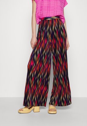 Kobieta nosząca szerokie plisowane spodnie z wielokolorowym abstrakcyjnym wzorem, różowy kraciasty top i beżowe sandały na platformie.