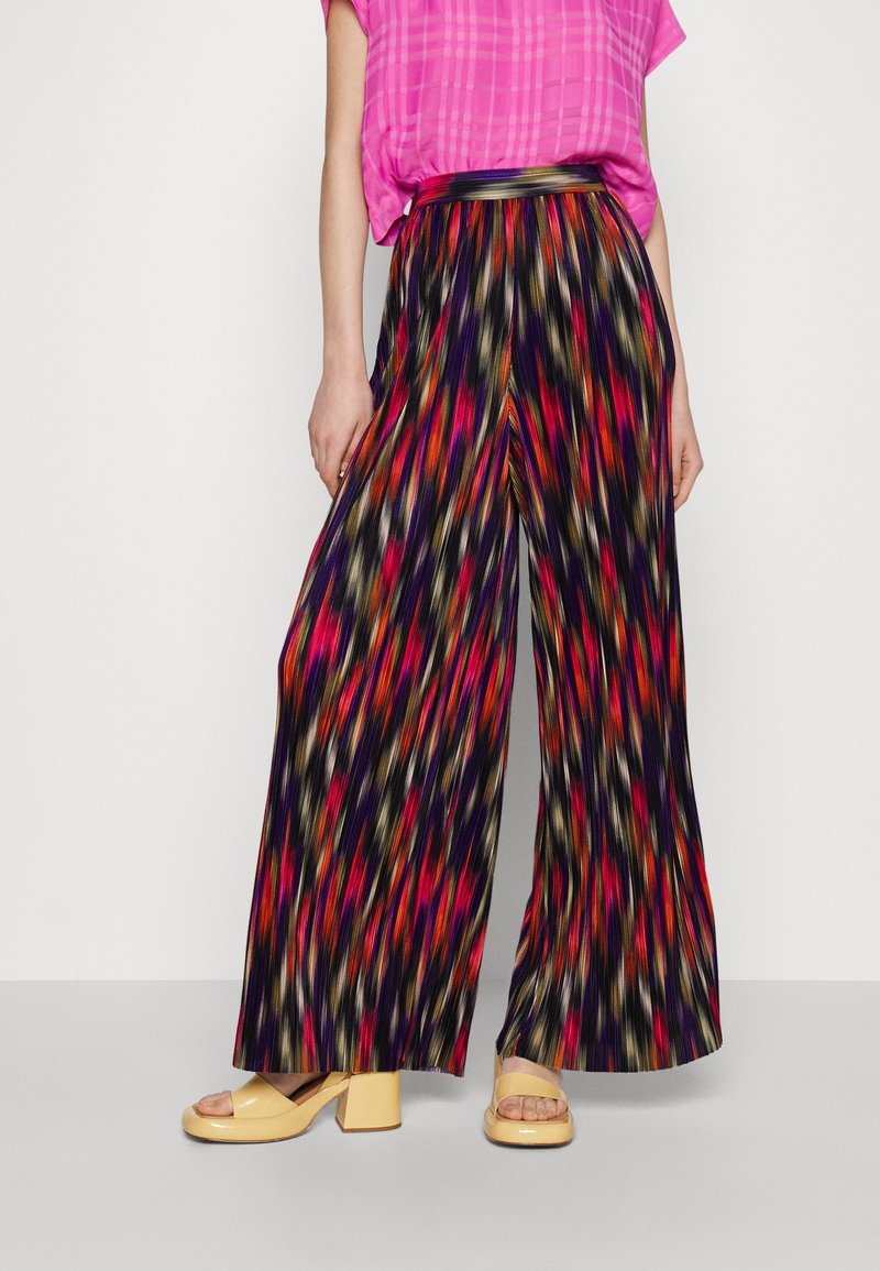 Femme portant un pantalon plissé à jambes larges avec un motif abstrait multicolore, un haut à carreaux rose et des sandales compensées beiges.