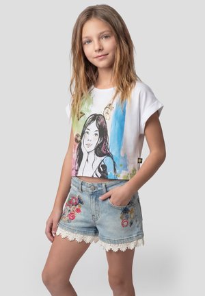 Niña joven con cabello largo, vistiendo una camiseta corta gráfica de colores y shorts de mezclilla con bordado floral y detalles de encaje, de pie con la mano en el bolsillo.