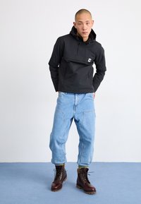 Giacca nera con cappuccio, tasca frontale, indossata con jeans larghi blu chiaro e stivali brown con lacci e suola testurizzata. Sfondo semplice.