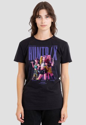 Woman wearing black T-shirt featuring three animated female characters with colorful outfits and the text "HUNTR/X" and names Zoey, Rumi, Mira.
