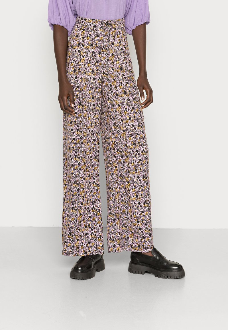 Calças largas florais com um padrão roxo, amarelo e preto, apresentando uma cintura alta e um corte descontraído, acompanhadas de sapatos plataforma pretos.