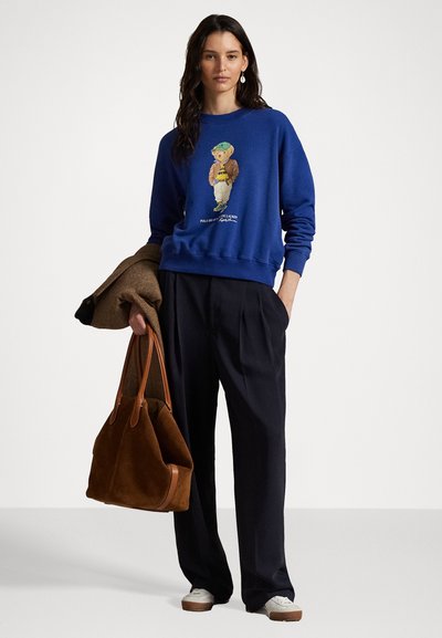 Blauwe sweatshirt met een beerprint, gecombineerd met donkere geplooide broeken en een bruine suède tote bag. Witte sneakers maken de outfit af.