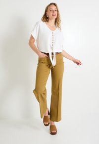 Top blanco de mangas cortas con escote en V y detalle de botones, combinado con pantalones anchos mostaza y sandalias marrones de tacón cuadrado.