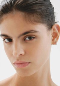 Primer plano de una persona con piel suave, cabello oscuro peinado hacia atrás, que lleva un pequeño aro de plata en la oreja izquierda. Iluminación natural suave.