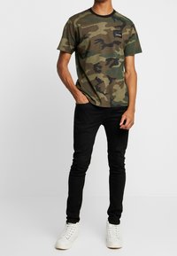 Camiseta de camuflaje en tonos verdes y marrones con mangas cortas y cuello redondo; combinada con jeans skinny negros y zapatillas blancas.