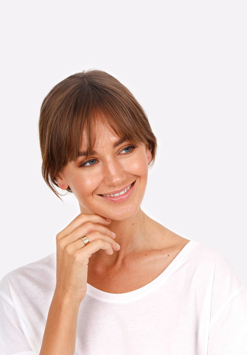 Gold ring with textured design worn on the left hand. Model wearing a white, wide-neck, short-sleeve shirt with a smooth texture. Light background.