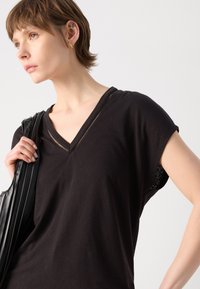 Top nero a scollo a V con maniche corte, caratterizzato da un tessuto testurizzato con dettagli a intaglio lungo il décolleté, abbinato a una borsa nera.