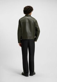 Giacca in pelle verde scuro con una texture liscia, caratterizzata da un colletto rialzato e un design snello, abbinata a pantaloni neri.