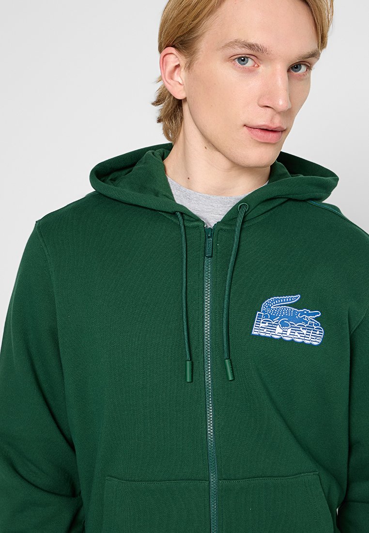 Sweat à capuche zippé vert en tissu doux, avec une poche avant, des cordons de serrage ajustables et un logo brodé bleu sur la poitrine.