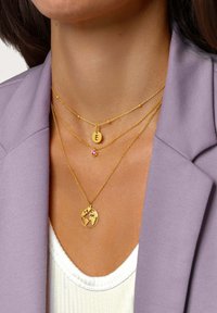 Colliers en métal doré avec un pendentif rond représentant des continents, un petit accent de pierre précieuse et un charm avec une initiale. Porté avec un blazer lavande.