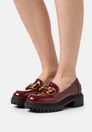 Mocassins bordeaux brillants avec une semelle plateforme noire épaisse, présentant un accent décoratif en chaîne dorée sur le devant. Texture lisse.