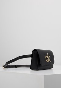 Černá kožená kabelka s texturovaným povrchem, která má zlatý CK logo na klopě, odnímatelný popruh a kompaktní tvar.