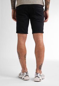Shorts in denim neri con risvolti, due tasche posteriori, indossati con scarpe da ginnastica bianche, tatuaggio visibile sulla gamba, mostrati da dietro.