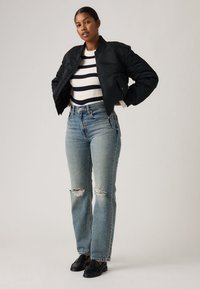 Donna in piedi con le mani sui fianchi, indossa una giacca bomber nera, un maglione a righe, jeans chiari strappati e mocassini neri su sfondo uniforme.