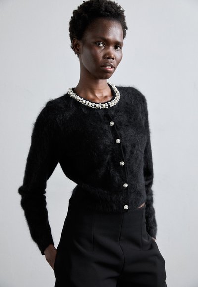 Schwarze, flauschige Strickjacke mit besticktem Kragen, mit einer Knopfleiste vorne und langen Ärmeln, kombiniert mit maßgeschneiderten schwarzen Hosen.