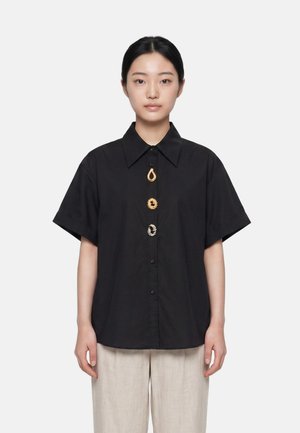 Donna che indossa una camicia nera a maniche corte con colletto oversize e tre grandi bottoni decorativi, abbinata a pantaloni beige chiaro.