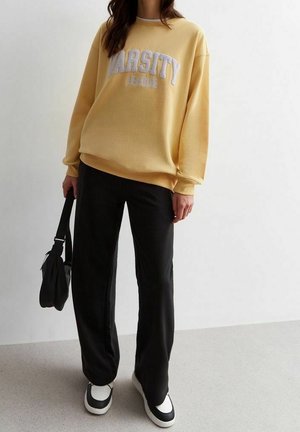 Osoba ubrana w żółtą bluzę „VARSITY LEAGUE”, czarne spodnie, czarno-białe trampki, trzymająca małą czarną torebkę na ramię.