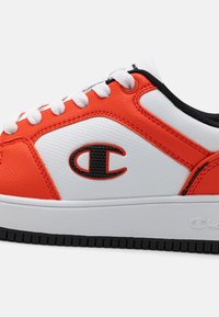 Baskets blanches et orange avec une finition texturée, présentant des accents et un logo noirs, un col rembourré et des lacets blancs plats sur une semelle noire.