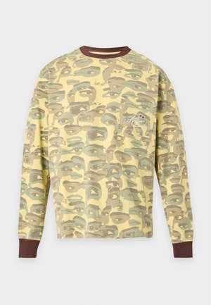 Homme debout portant une chemise à manches longues jaune et verte avec un motif camouflage et un pantalon large assorti ainsi que des baskets noires et blanches.