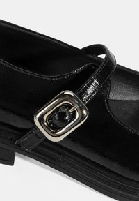 Schwarzer Lacklederschuh mit abgerundetem Zehenbereich, ausgestattet mit einem schmalen Knöchelriemen und einem quadratischen silbernen Schnallenakzent. Strukturiertes Oberflächendetail.